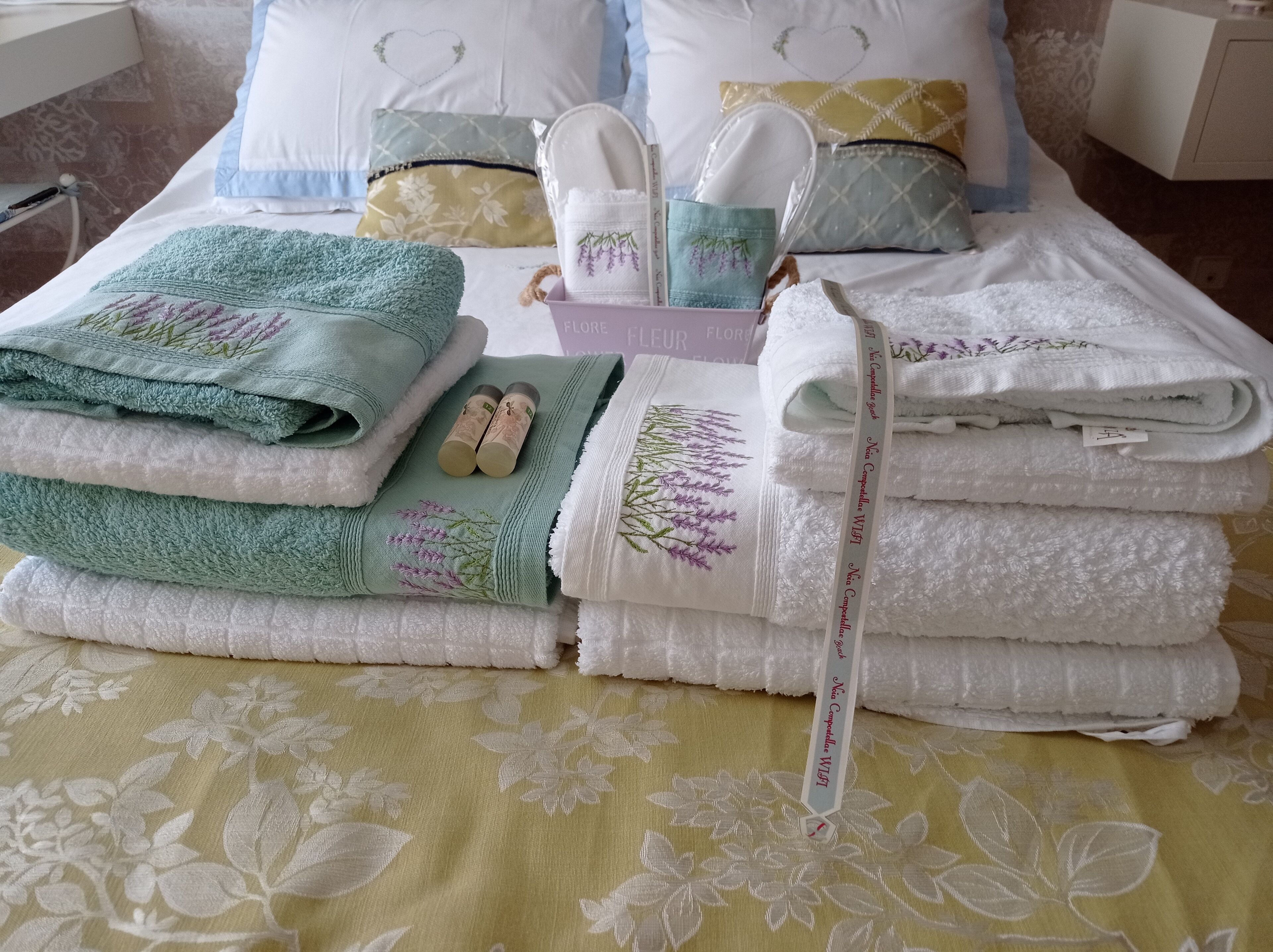 Una cama bien hecha con toallas dobladas y un par de pantuflas, un almohadón con diseño de corazón y un libro titulado "Fleur".