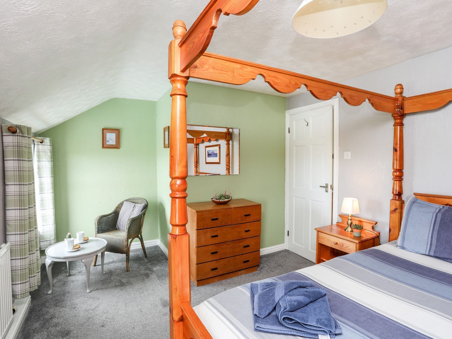 A four-poster bed, a wooden dresser, a chair, a small table, and a door in a bedroom.