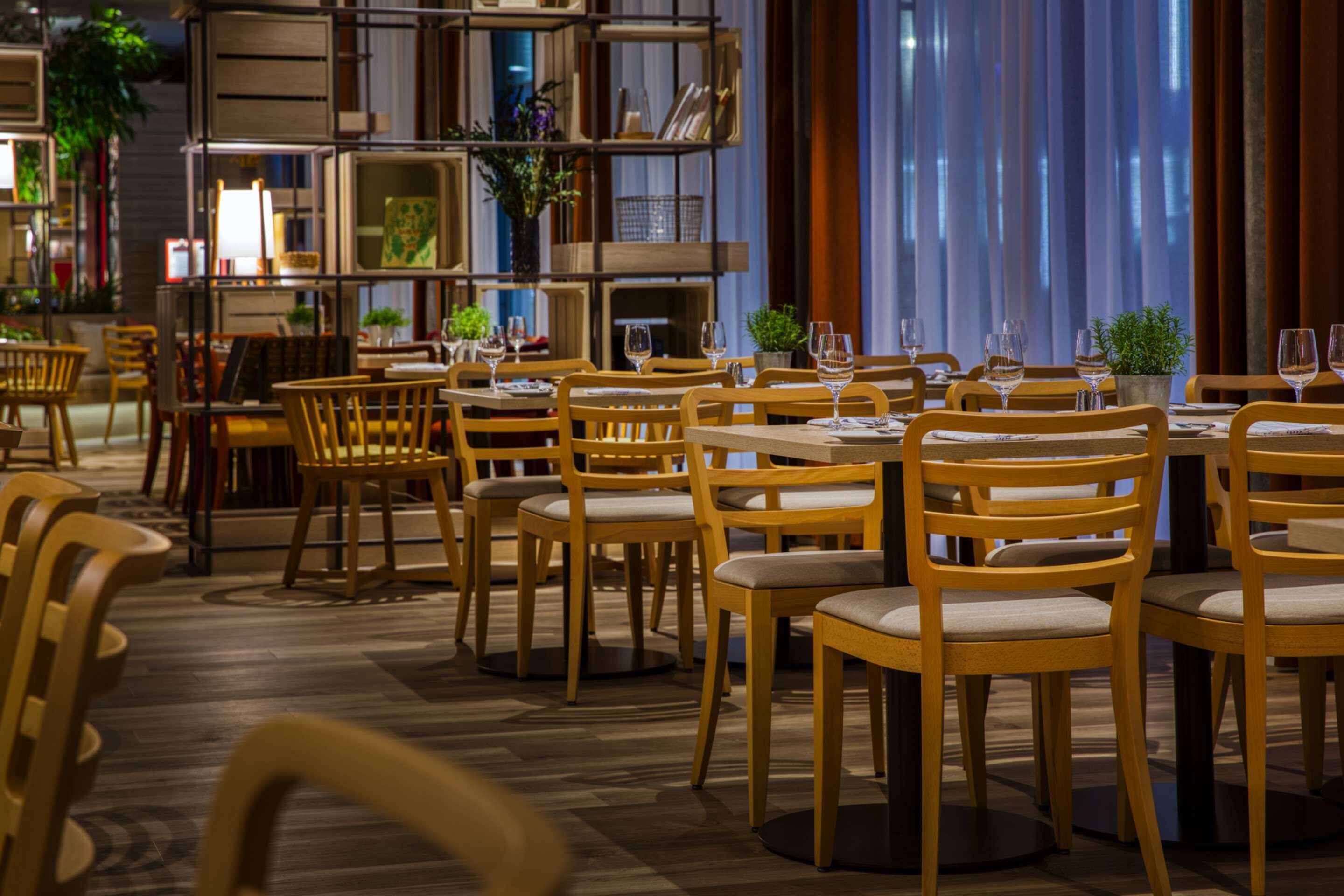 A restaurant with wooden chairs and tables, a bookshelf, and a large window with curtains.