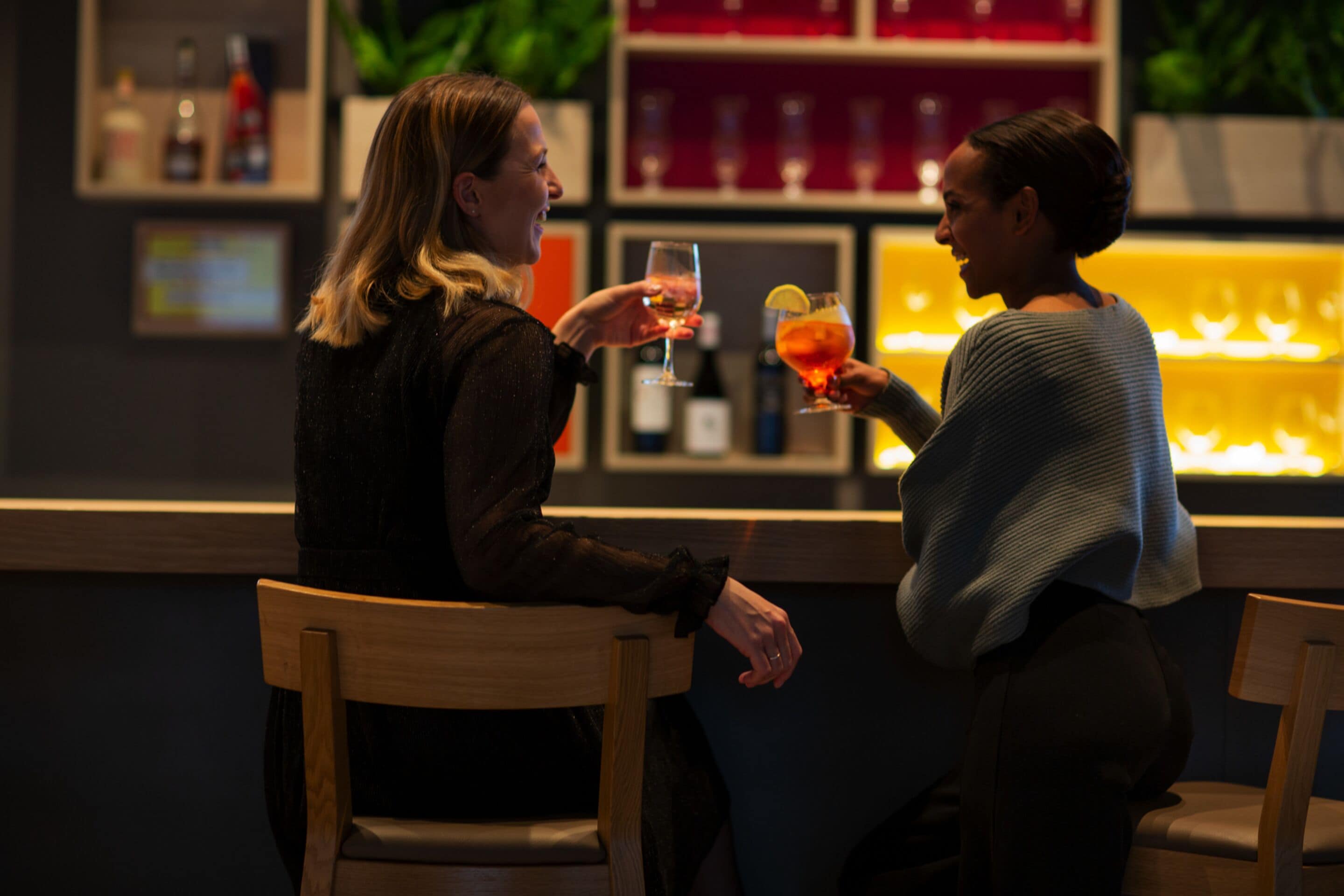 Dos personas sentadas en un bar, con una estantería llena de botellas y vasos al fondo.