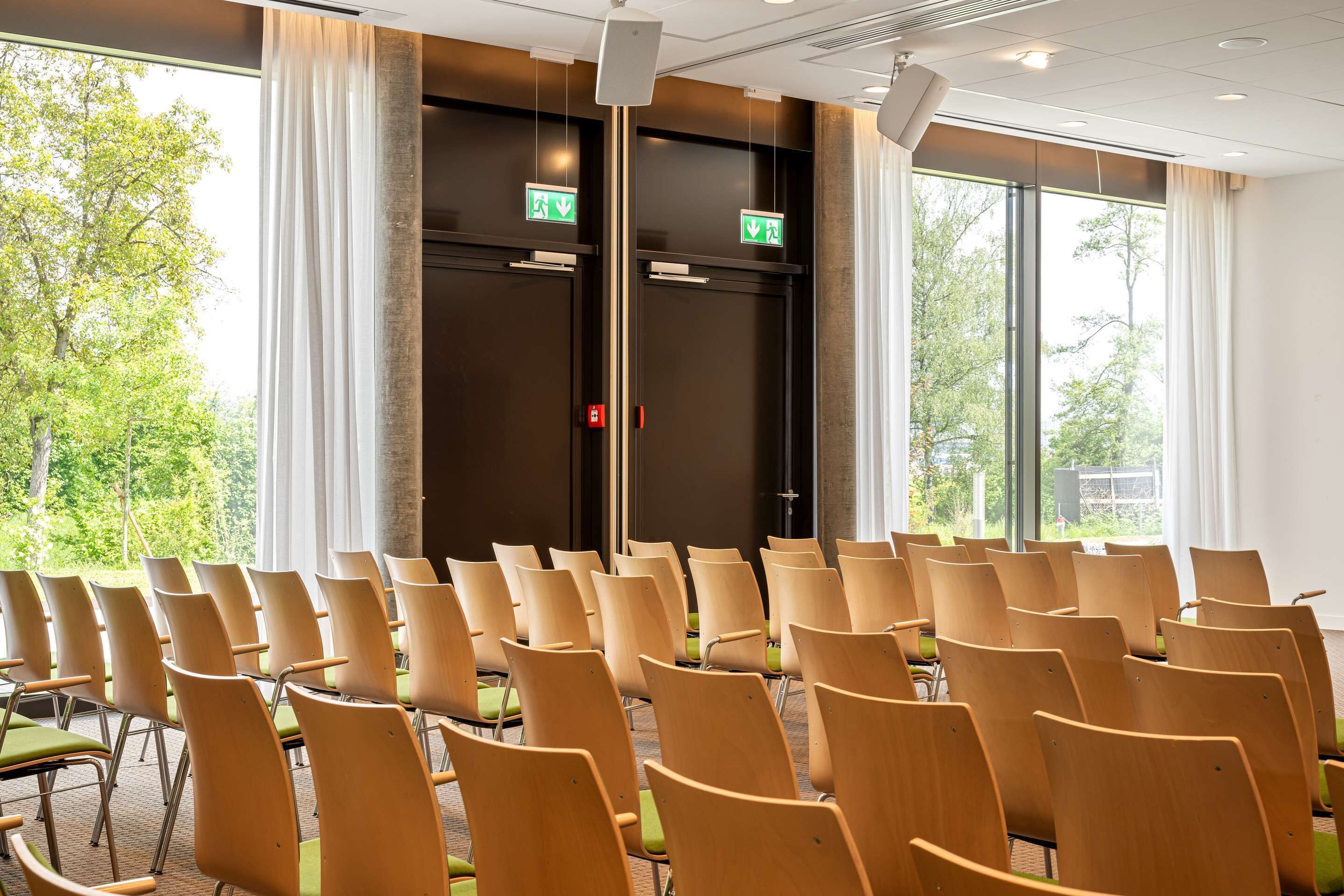 A conference room with rows of chairs, large windows, and two doors with emergency exit signs.