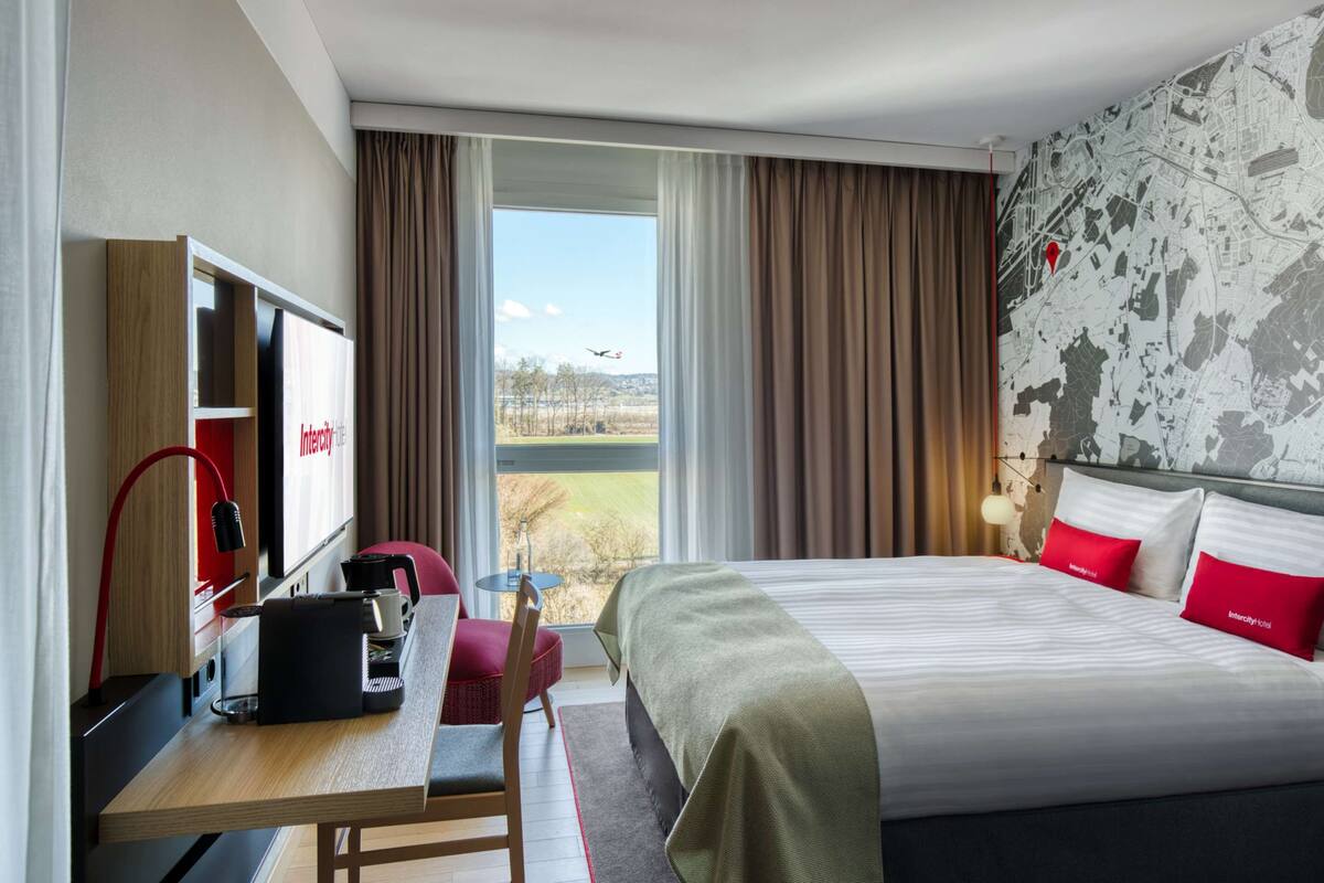 Habitación de hotel con cama, escritorio, silla, televisor y ventana con cortinas.