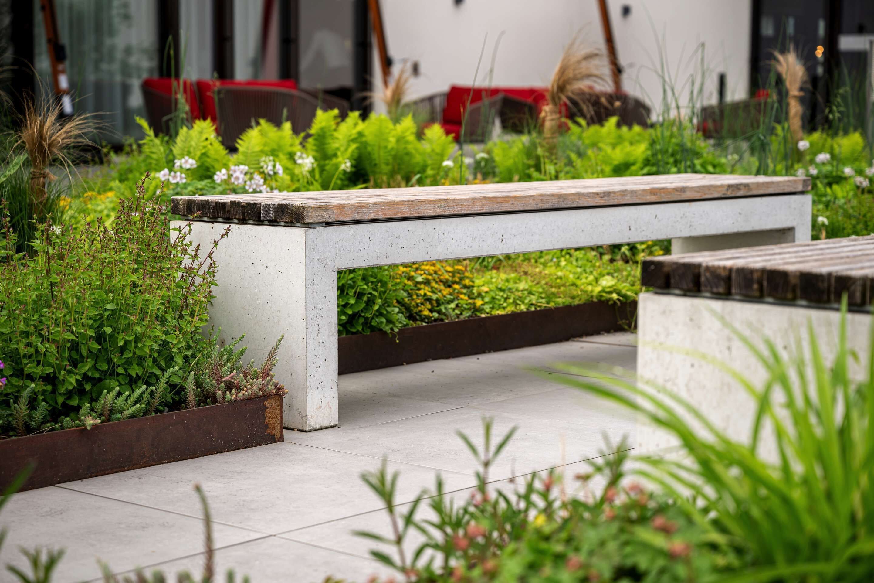 A white bench in a garden area with various plants and a concrete planter.