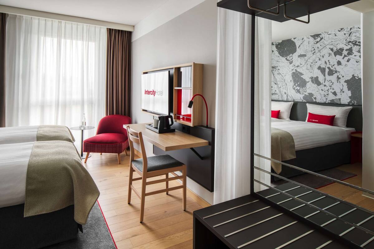 Habitación de hotel con dos camas, un escritorio con ordenador, una silla roja y un espejo grande.