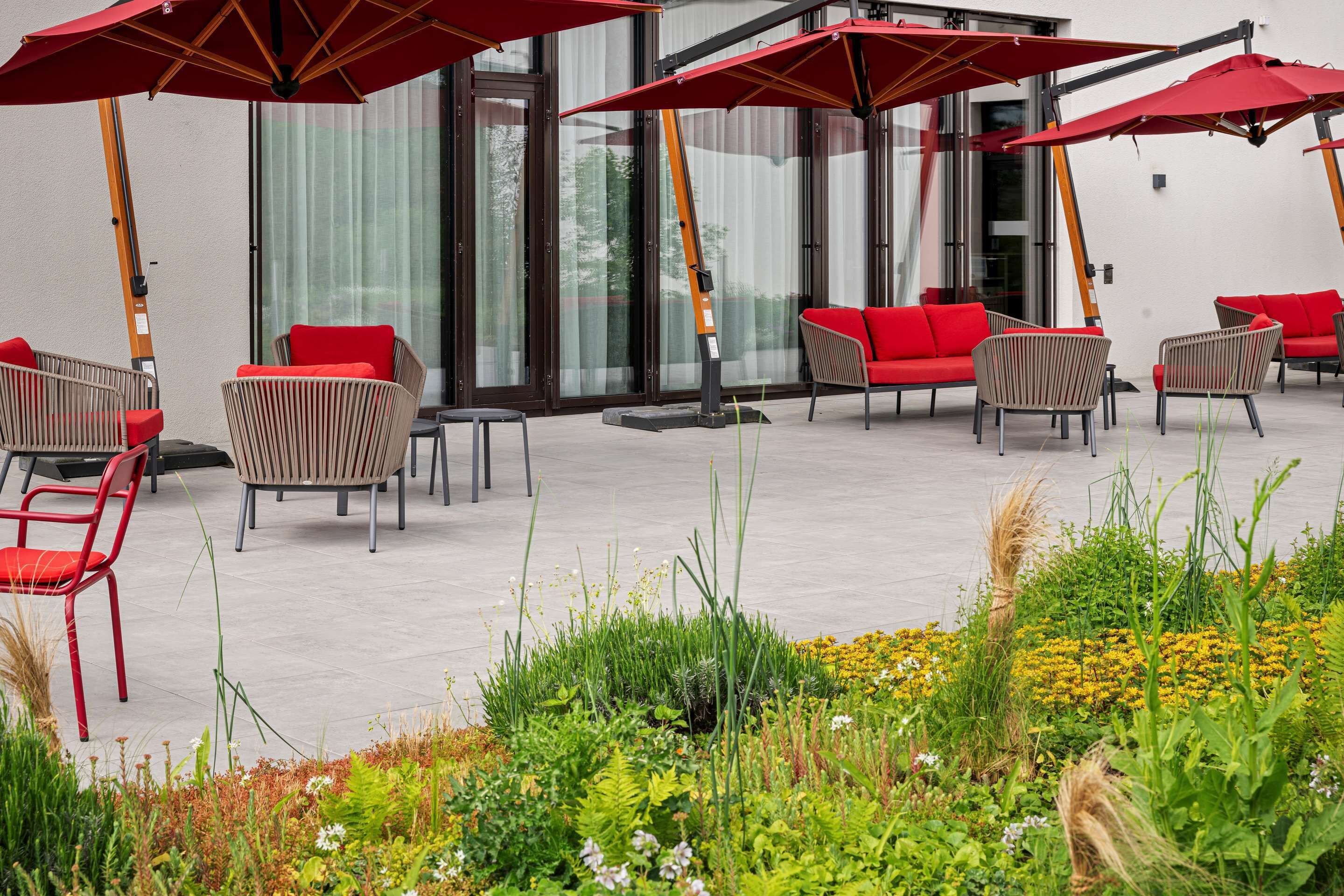 Terrassa exterior amb cadires vermelles i paraigües, un jardí amb diferents plantes, i un edifici amb grans finestres al fons.
