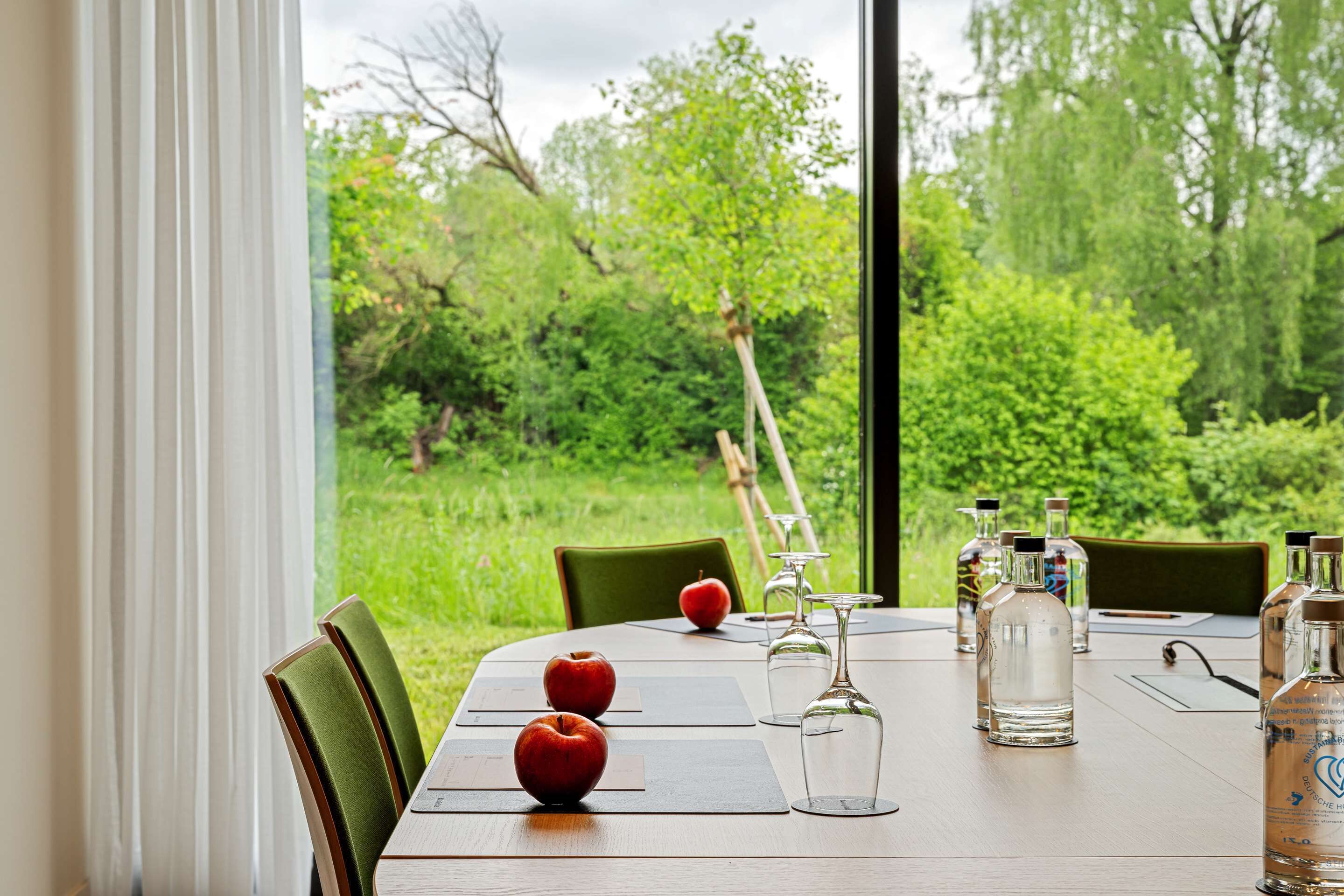 A dining table with three apples, wine glasses, and bottles, set against a backdrop of lush greenery.