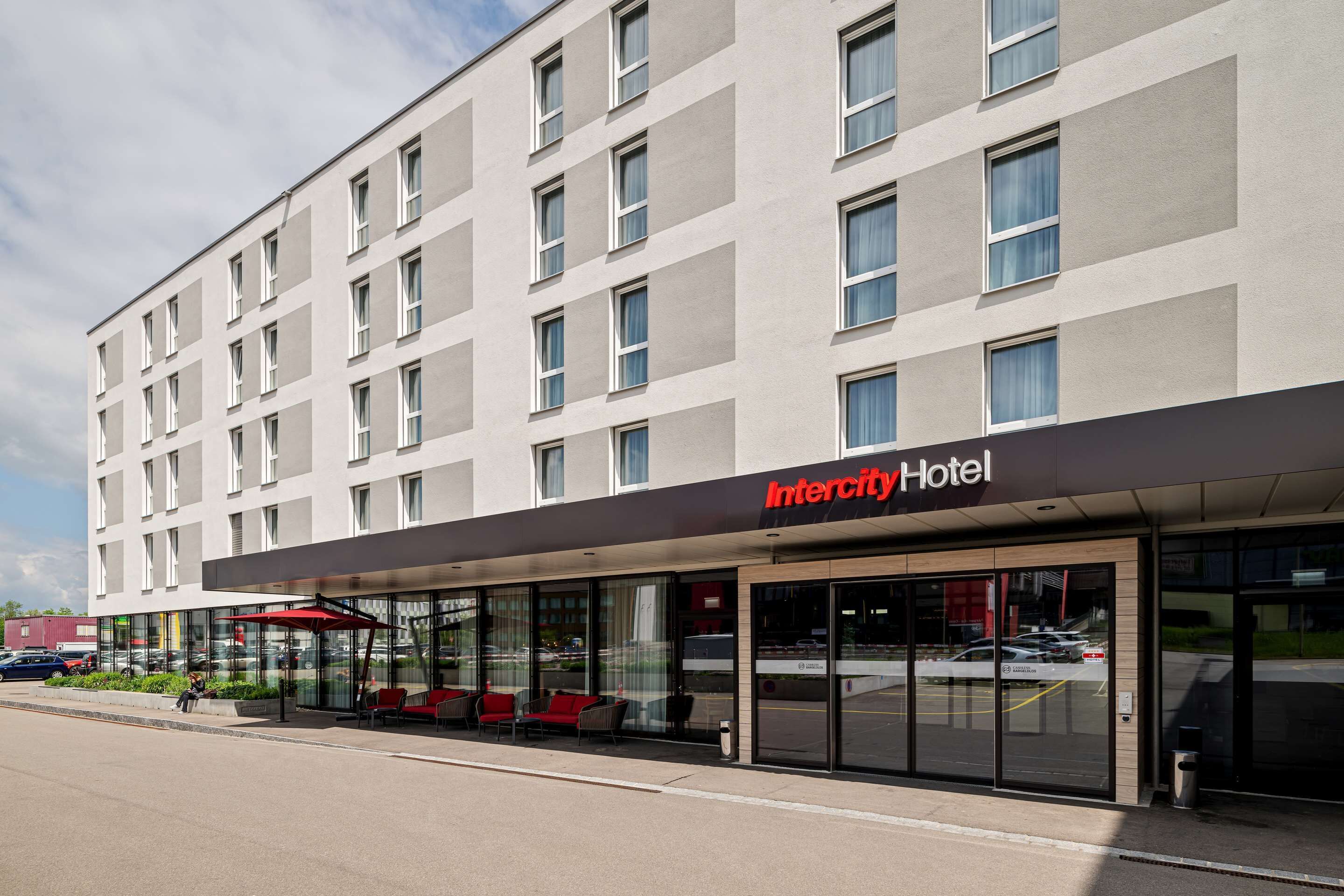 Ein modernes Intercity Hotel mit Glasfassade, Außenbereich und einem Parkplatz im Hintergrund.