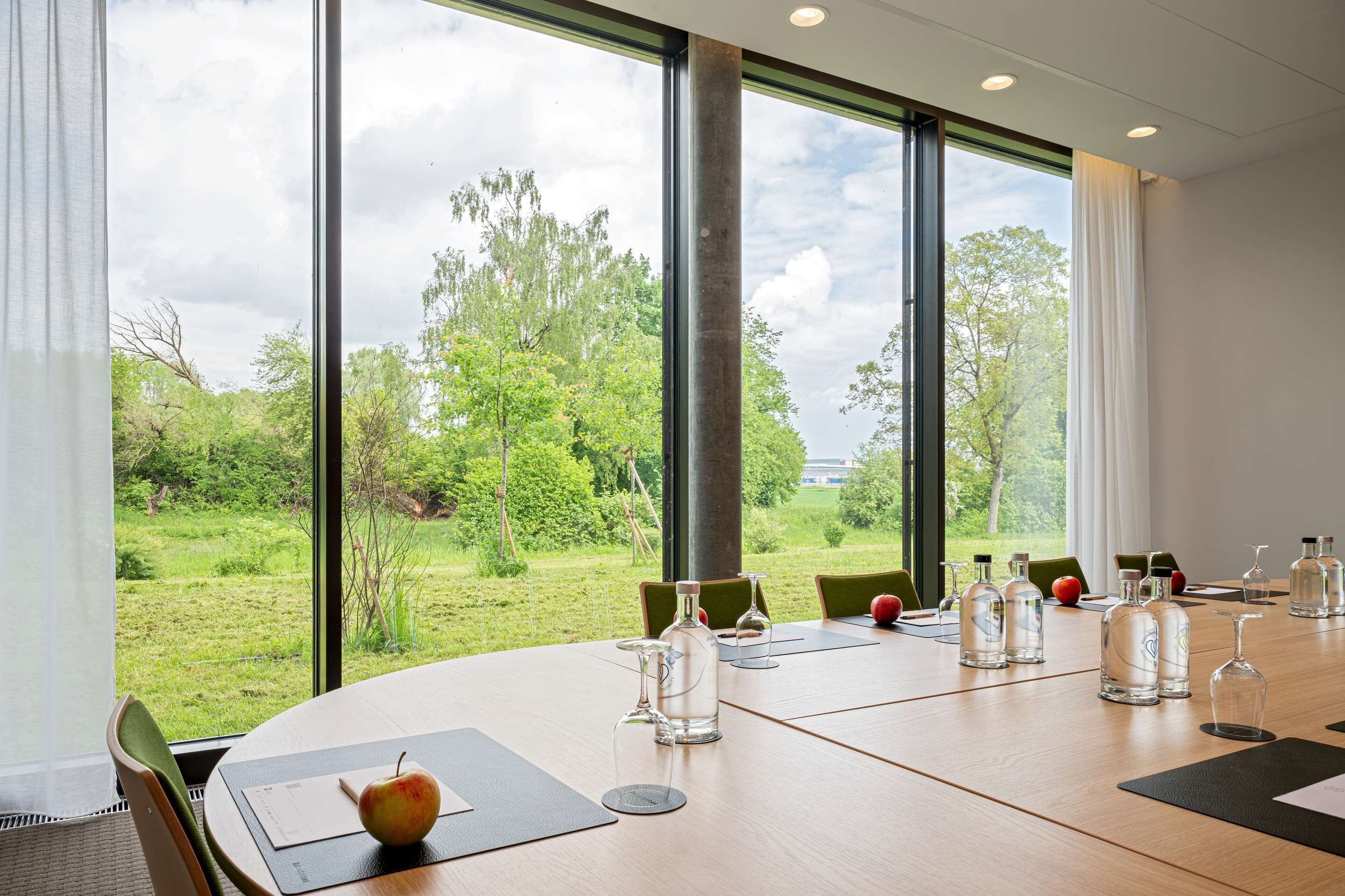 Un salón de conferencias con una gran mesa, sillas y vistas a un paisaje verde a través de amplios ventanales.