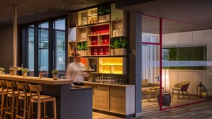 Een moderne bar met een bartender, houten barkrukken, glazenplanken en een uitzicht door een glazen deur op een zithoek met een bank en stoelen.