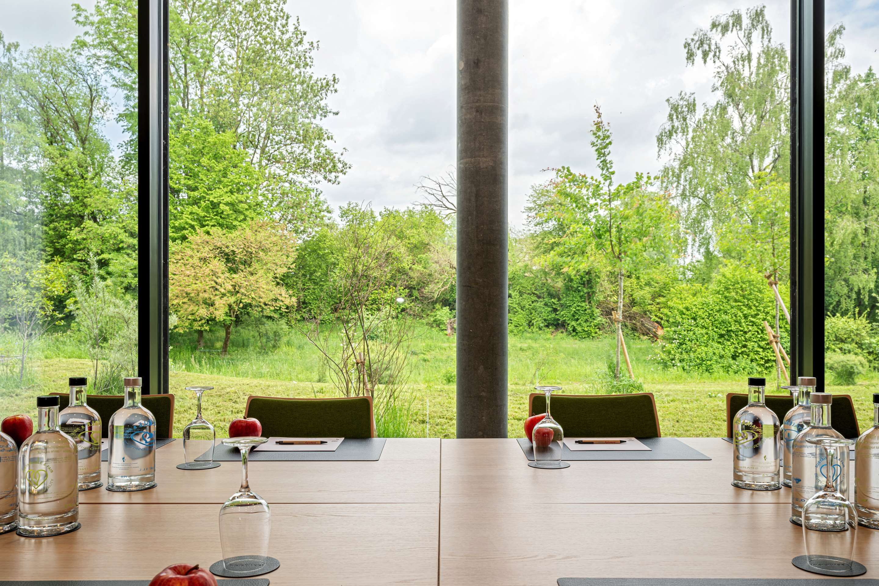 Mesa de comedor dispuesta con copas de vino, botellas y manzanas, con vistas a un paisaje verde exuberante a través de amplios ventanales.