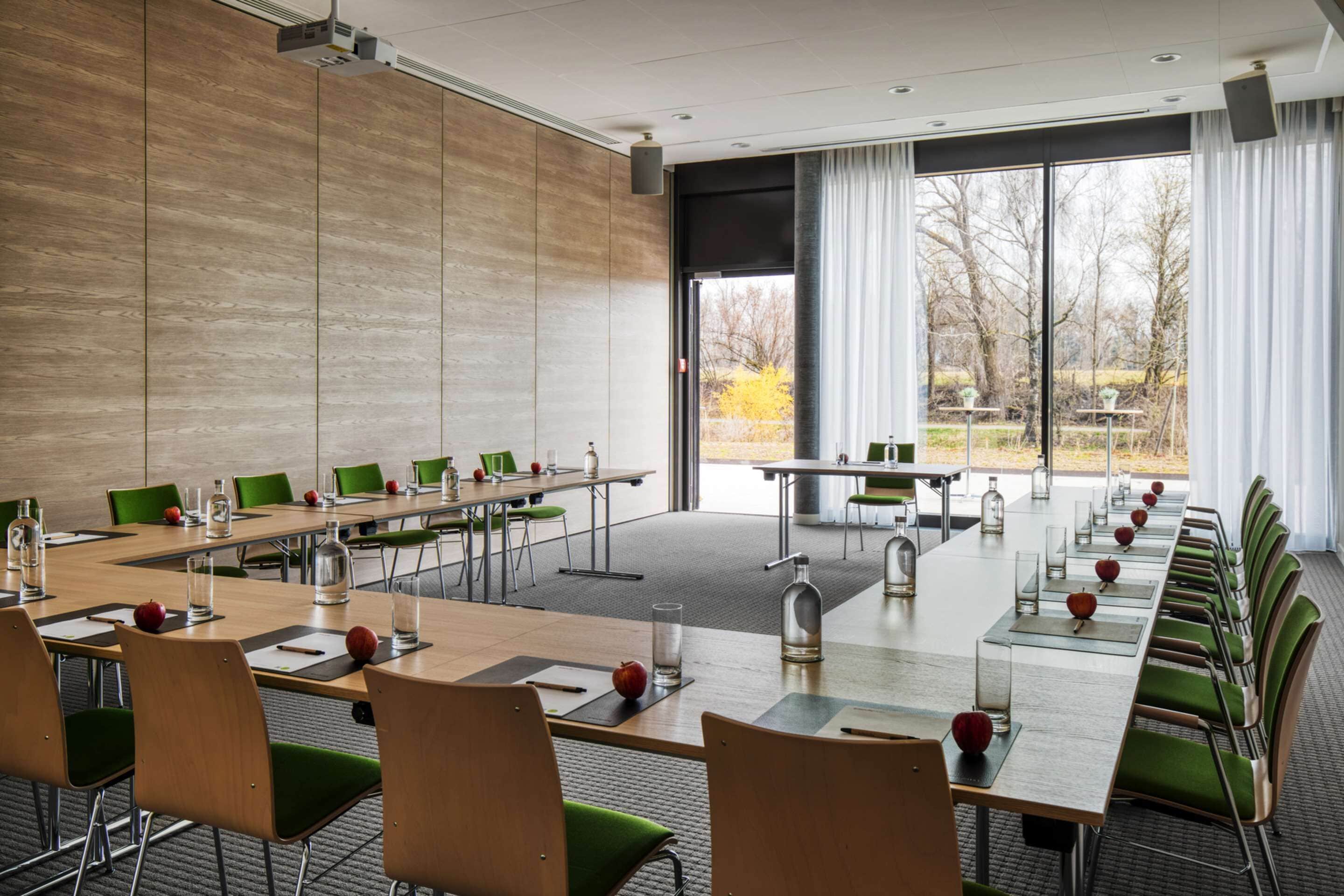 Ein Konferenzraum mit langen Tischen, grünen Stühlen und einem großen Fenster mit Blick auf Bäume.