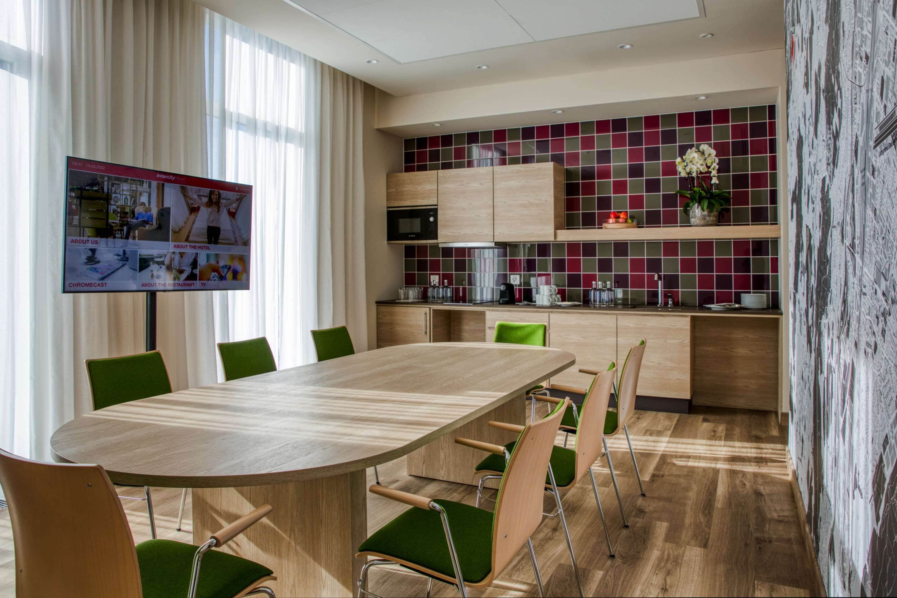 Una habitación de hotel moderna con una mesa de comedor de madera, sillas tapizadas en verde, un televisor de pantalla plana grande y una zona de cocina con baldosas rojas y verdes.