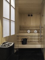A sauna with wooden benches, a black bucket with a wooden handle, and two clocks on the wall.