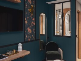 A room with a dark teal wall, a mounted TV, a decorative floral wallpaper, a mirror, a window with a view of a staircase, a dark armchair, and a wooden side table with a tray and glasses.