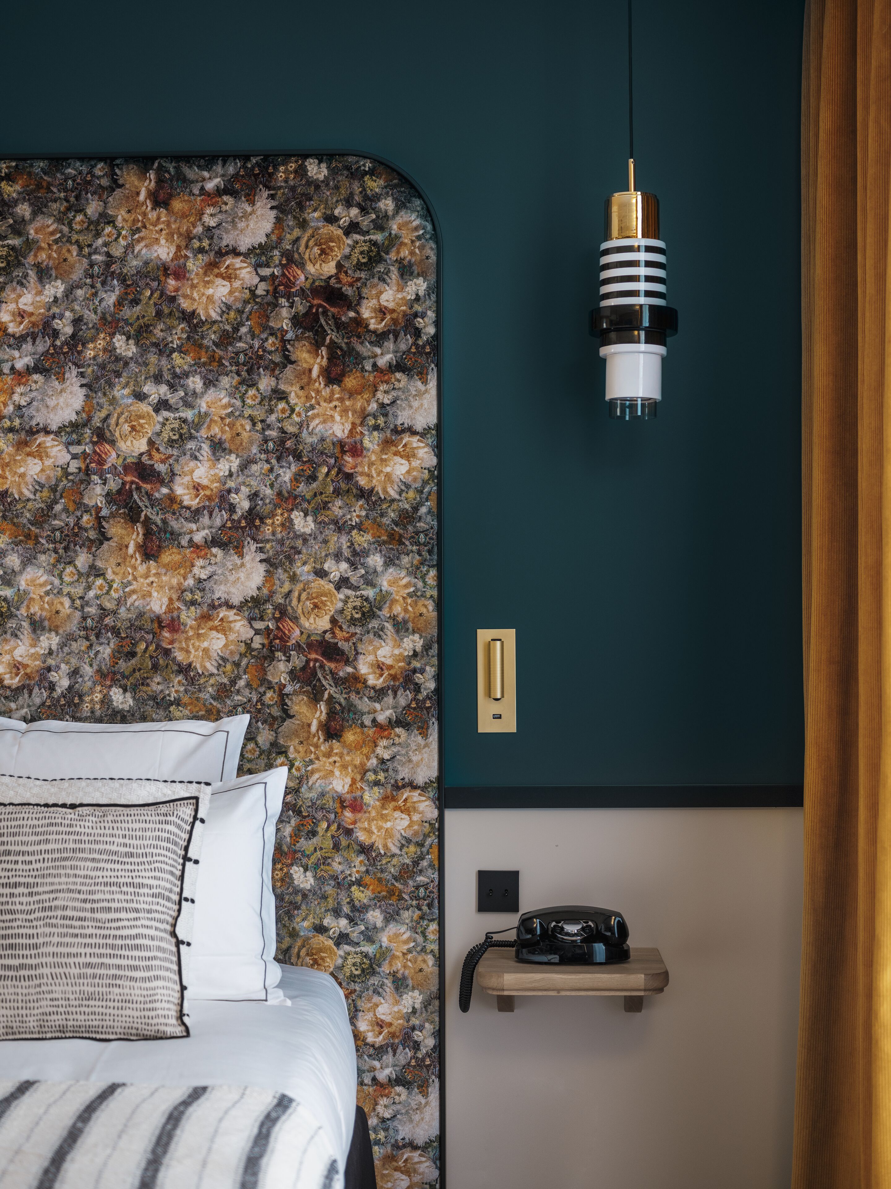 A bedroom with a floral wallpaper, a bed with white and striped bedding, a bedside table with a telephone, and a hanging light fixture.