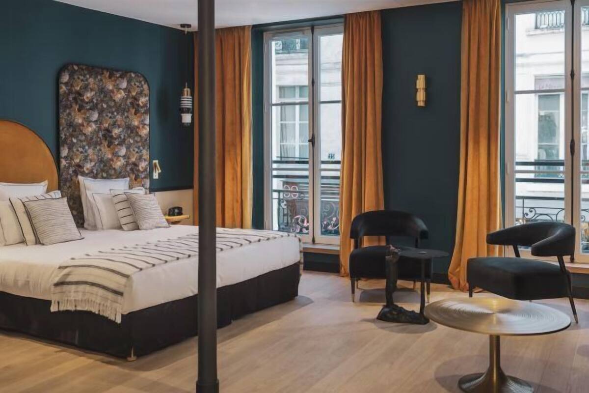 A hotel room with a bed, a large window with curtains, a chair, and a small table.