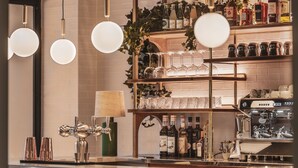 A bar area with wooden bar stools, a coffee machine, and shelves stocked with bottles and glasses. - Dandy Hotel  (Paris)
