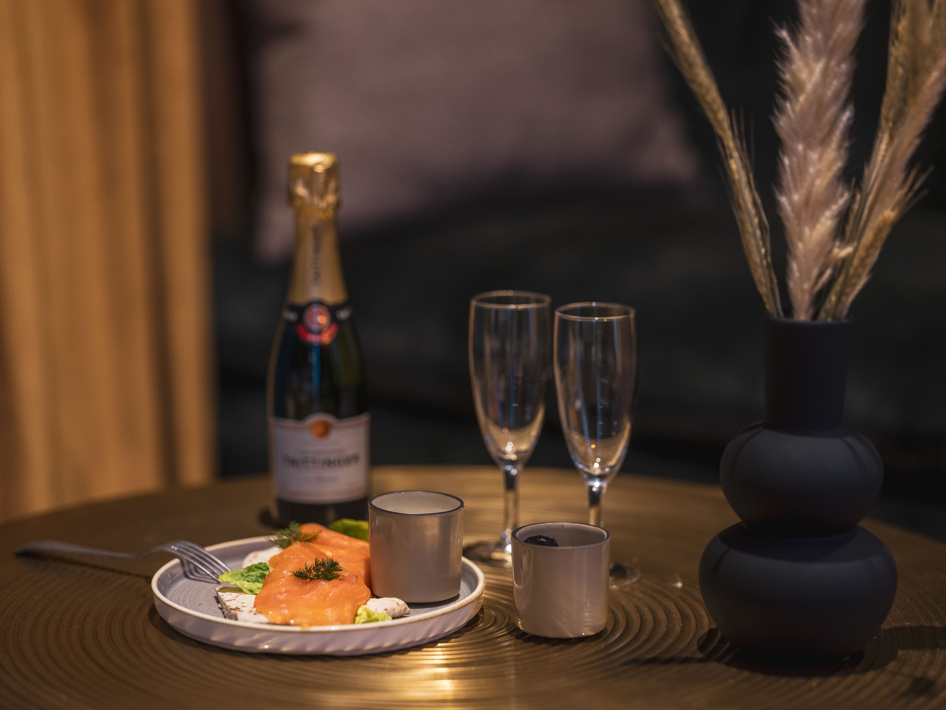 A table with a plate of food, a bottle of champagne, two glasses, and a vase with decorative elements.
