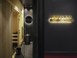 Un couloir avec un panneau indiquant la présence d’un sauna, une porte en bois, un miroir rond et une poubelle.
