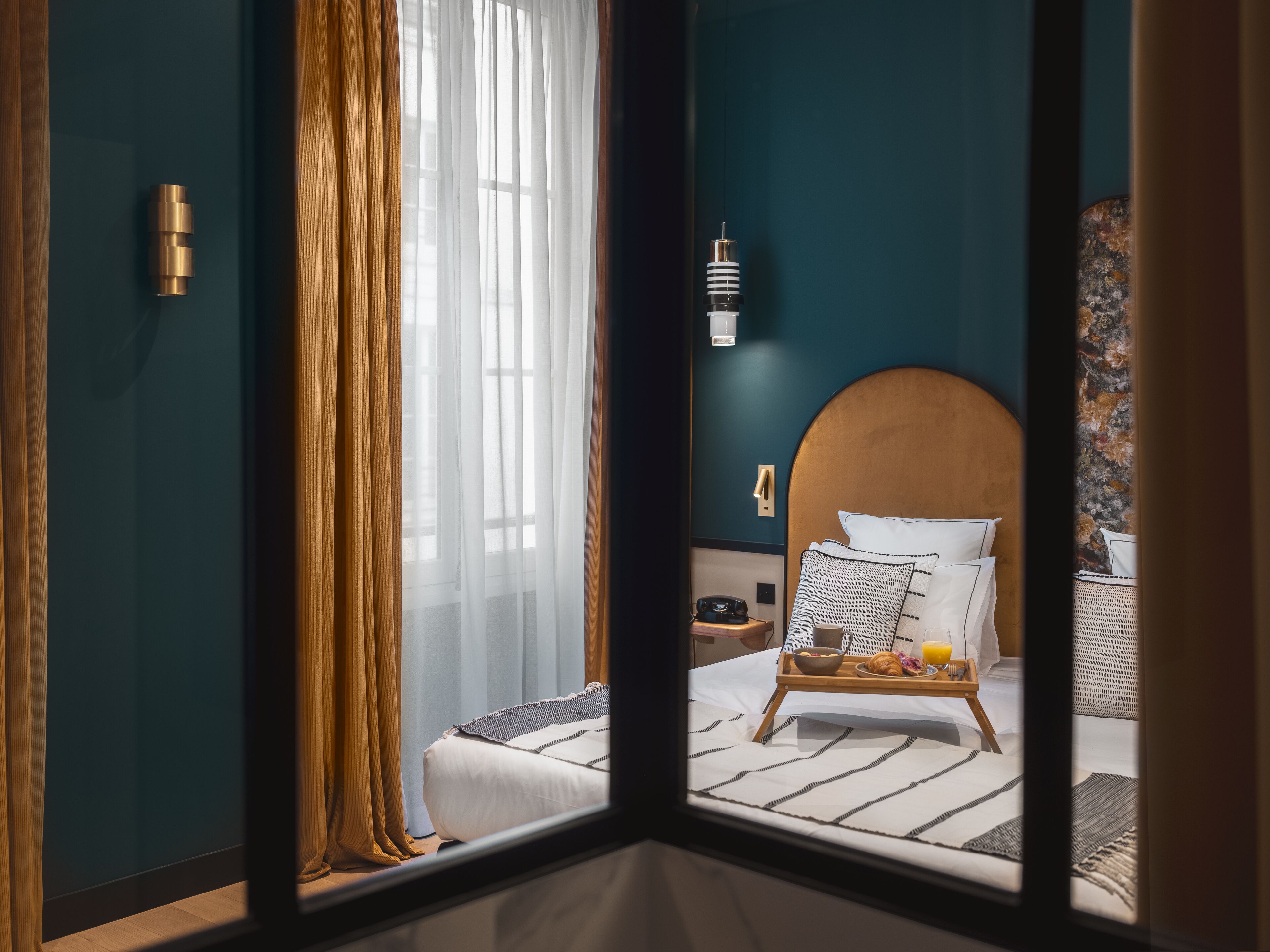 Una habitación de hotel con un ventanal grande, una cama con cabecera de madera, una mesita de noche con una lámpara y una bandeja con alimentos para el desayuno.
