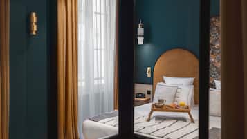 Una habitación de hotel con un ventanal grande, una cama con cabecera de madera, una mesita de noche con una lámpara y una bandeja con alimentos para el desayuno.