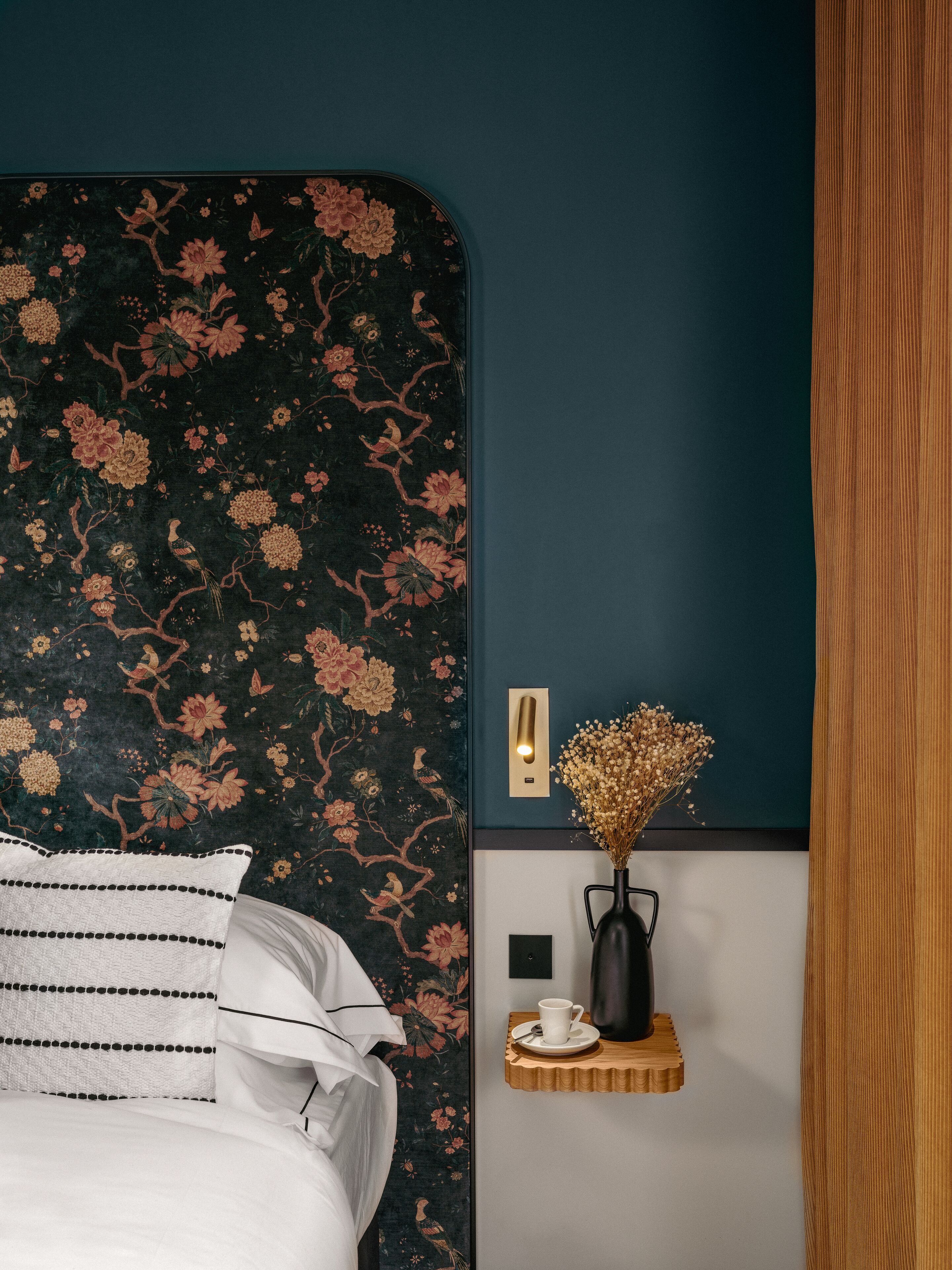 Un dormitorio con un cabecero de flores oscuras, una mesita de noche con una taza y un plato, y una lámpara de pared.