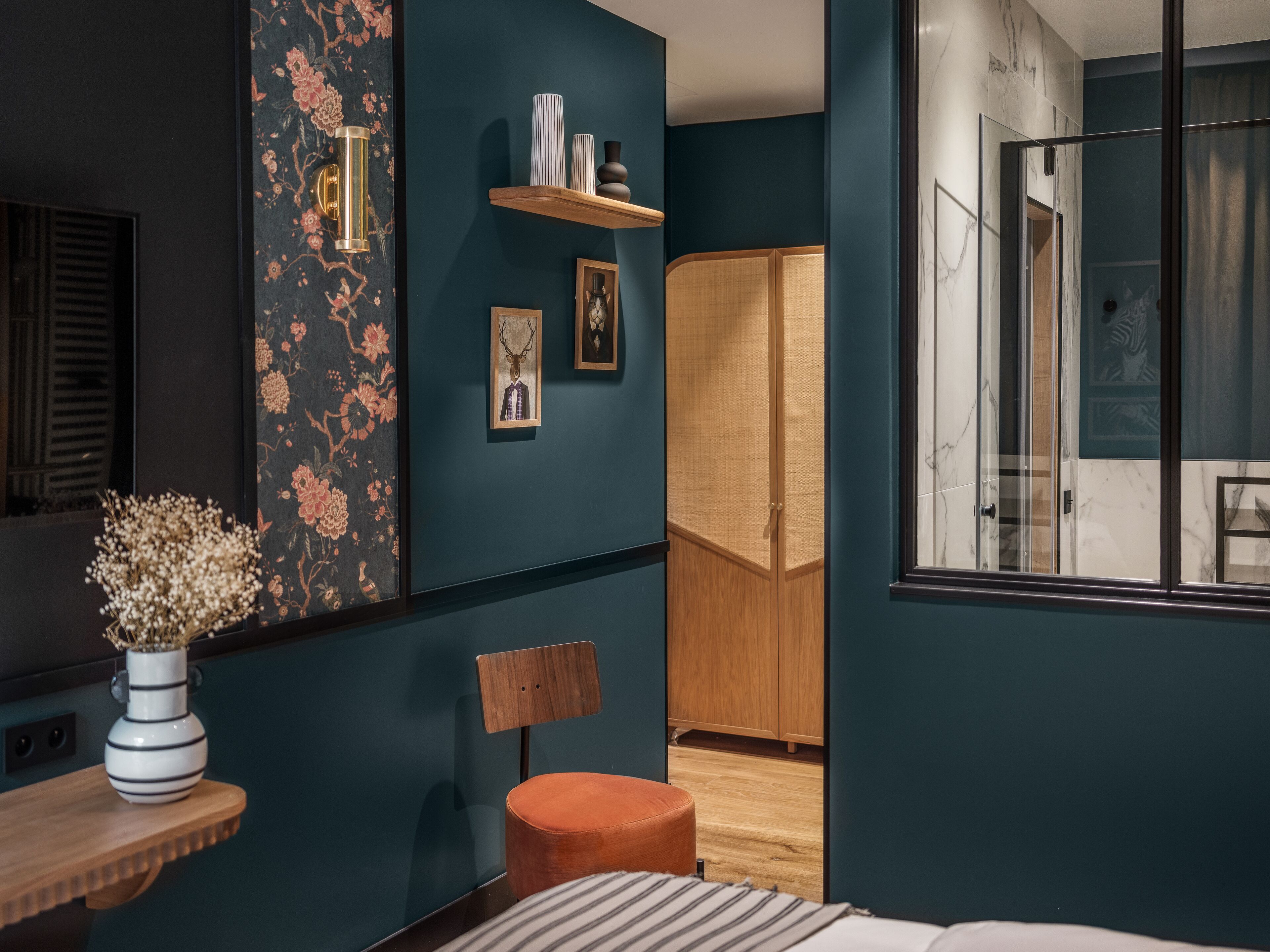 A modern bedroom with a large window, a wooden wardrobe, a small orange ottoman, and a wall-mounted shelf with decorative items.