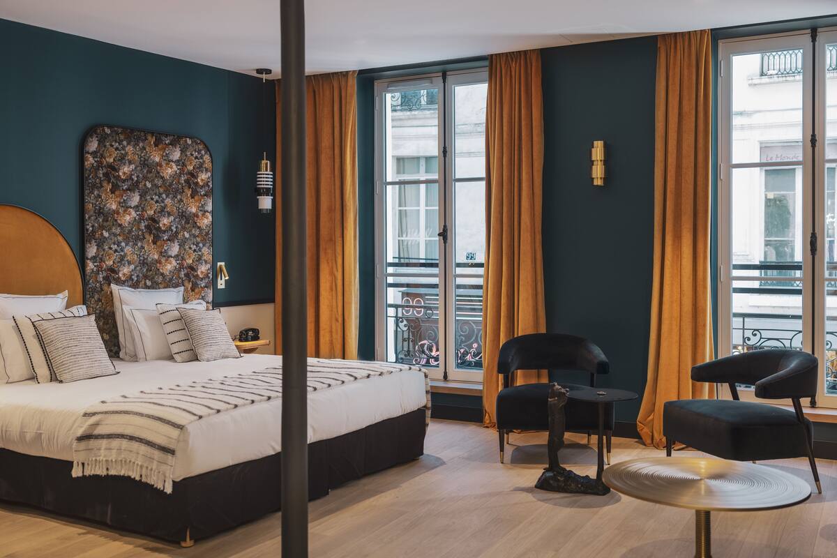 A modern hotel room with a large bed, two black chairs, a small round table, and a large window with curtains.