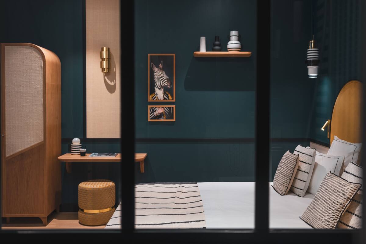 A modern bedroom with a bed, pillows, a bedside table, a small stool, a wall-mounted shelf with decorative items, and a framed picture.