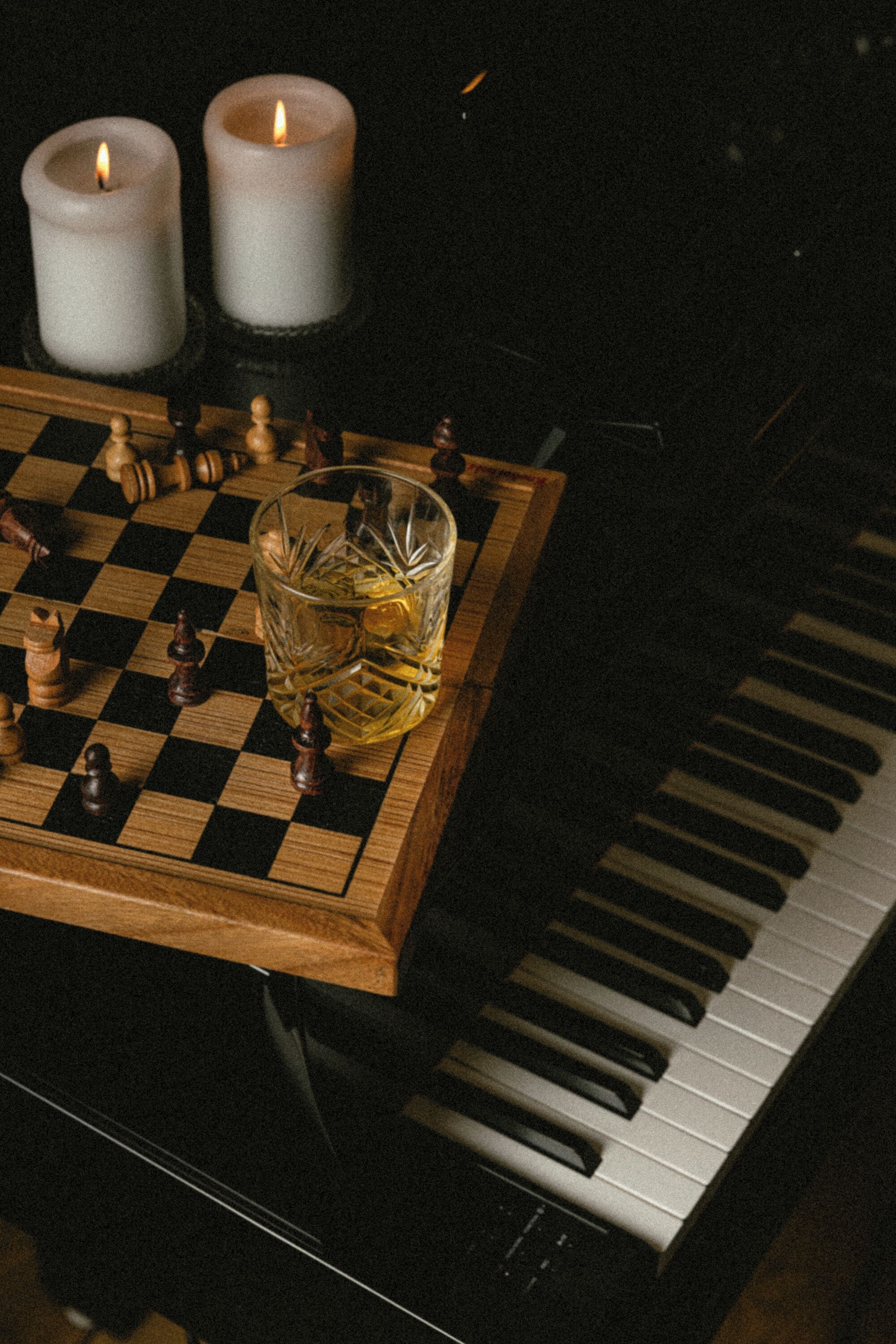 A checkered table with chess pieces, two lit candles, and a glass of amber liquid.