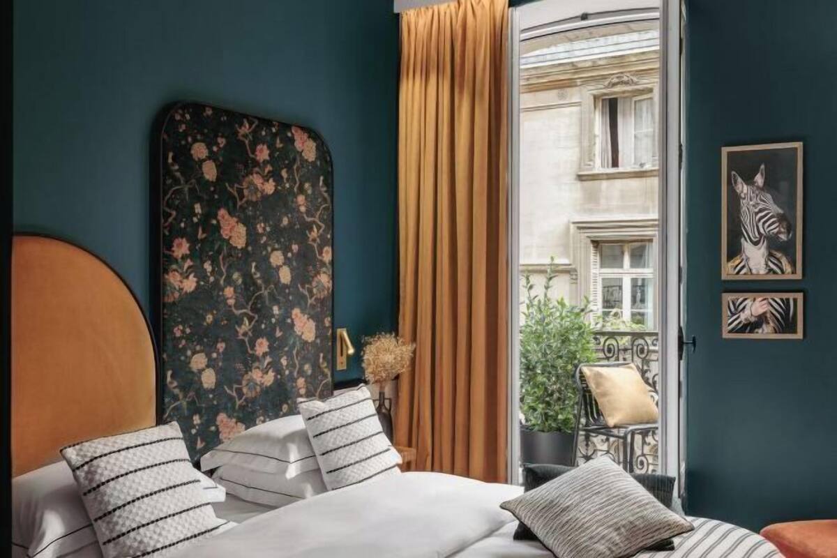 A bedroom with a bed, a headboard, a window with curtains, and a view of a building outside.