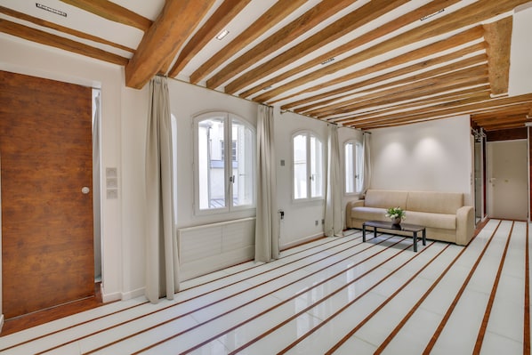 A spacious room with a high ceiling, wooden beams, large windows, a beige sofa, and a tiled floor.