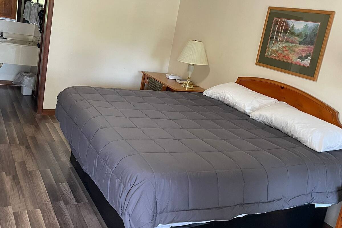 A bedroom with a bed, a nightstand, a lamp, a framed picture, and a wooden floor.