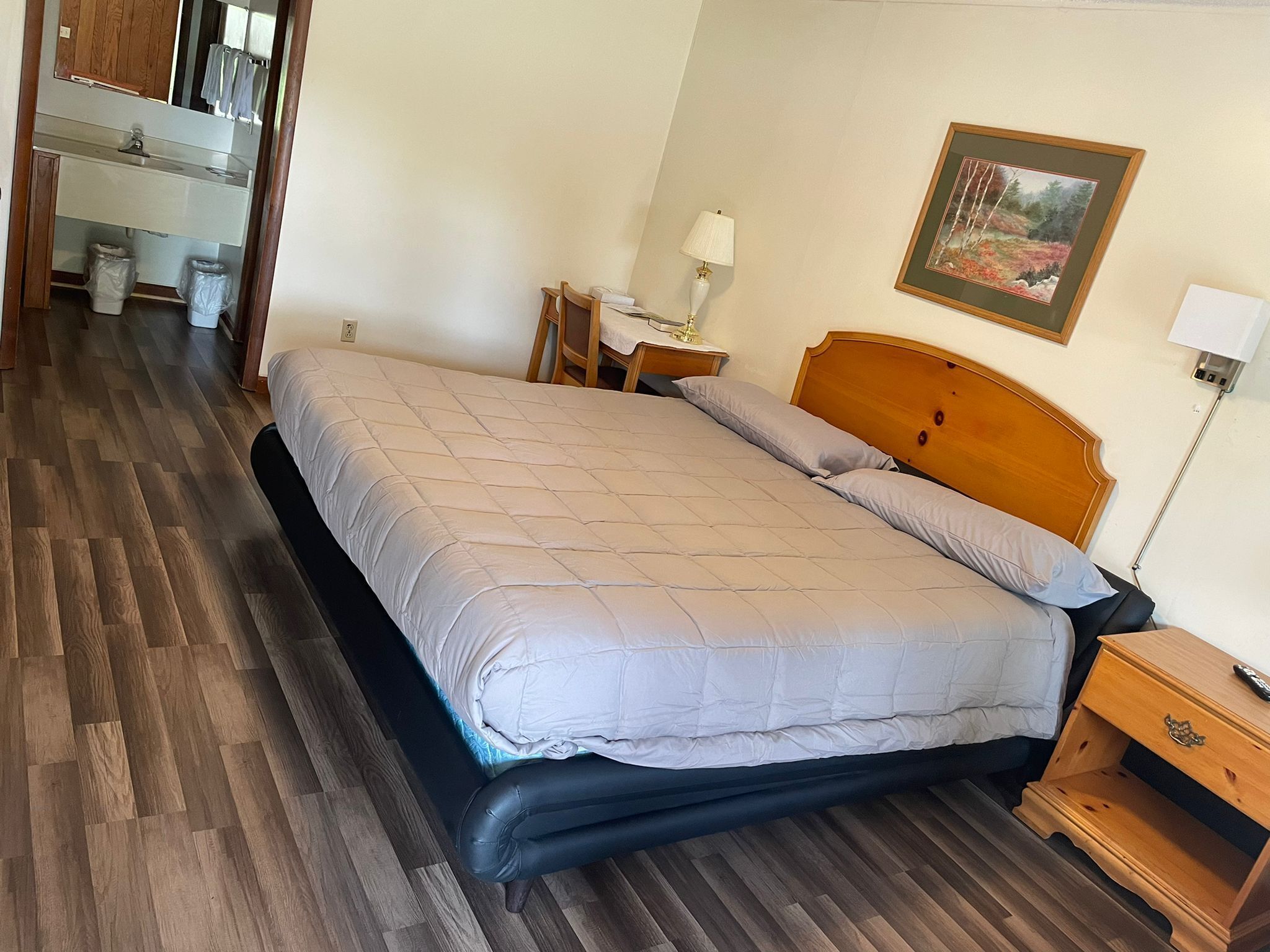 A bedroom with a bed, wooden headboard, bedside tables, a lamp, a framed picture on the wall, and a bathroom visible in the background.
