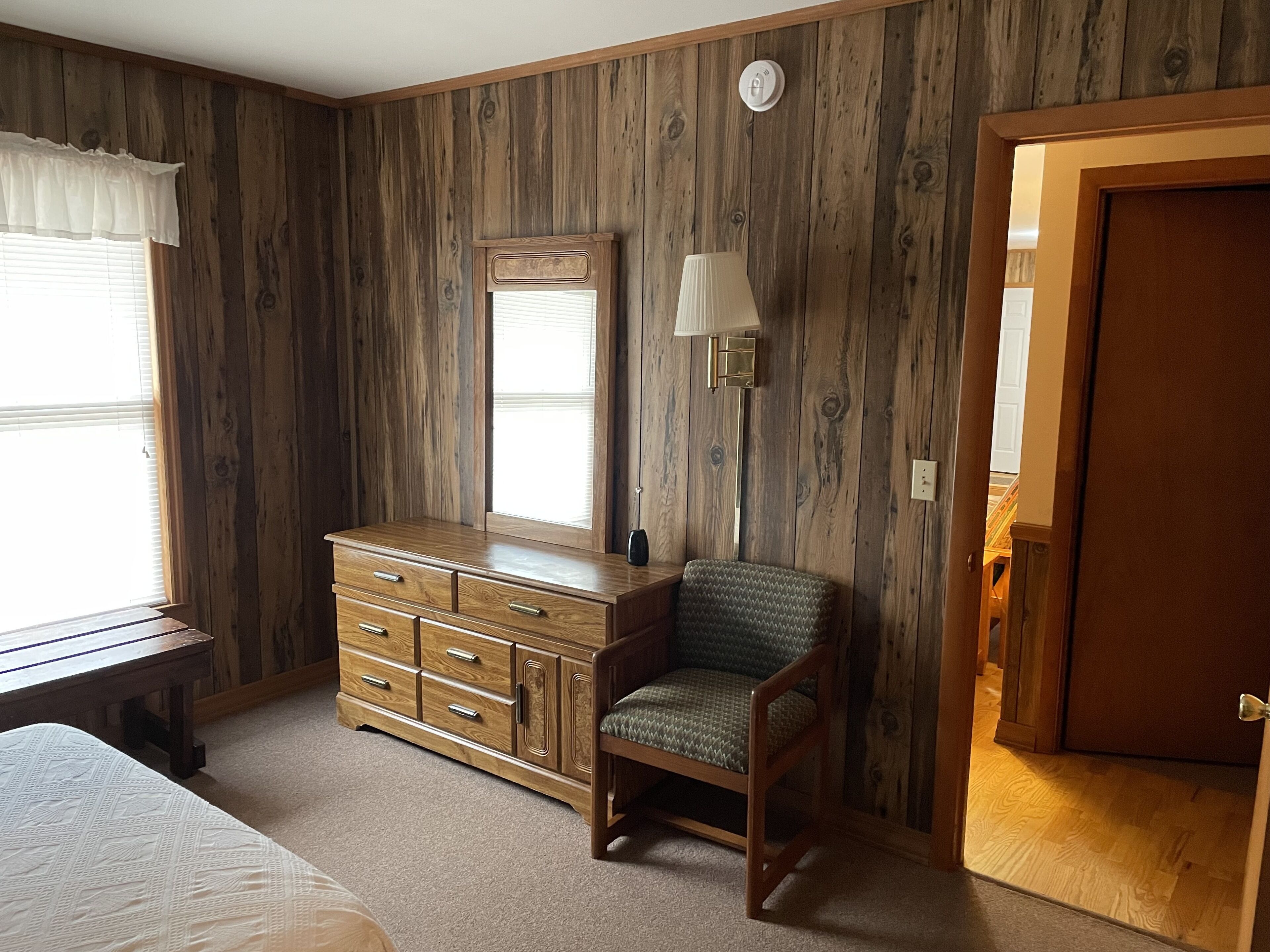 A bedroom with wooden panel walls, a wooden dresser, a chair, a bedside table, a lamp, and a door.