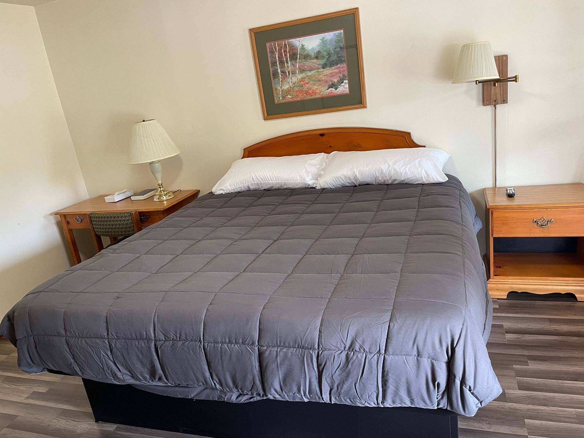 A neatly made bed with a grey quilt, a wooden headboard, a bedside table with a lamp, and a nightstand with a remote control.