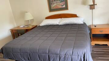A neatly made bed with a grey quilt, a wooden headboard, a bedside table with a lamp, and a nightstand with a remote control.