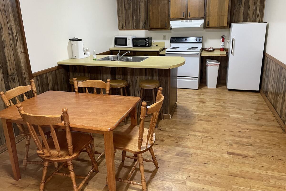A compact kitchen with wooden cabinets, a dining table with chairs, and basic appliances including a microwave, stove, and refrigerator.