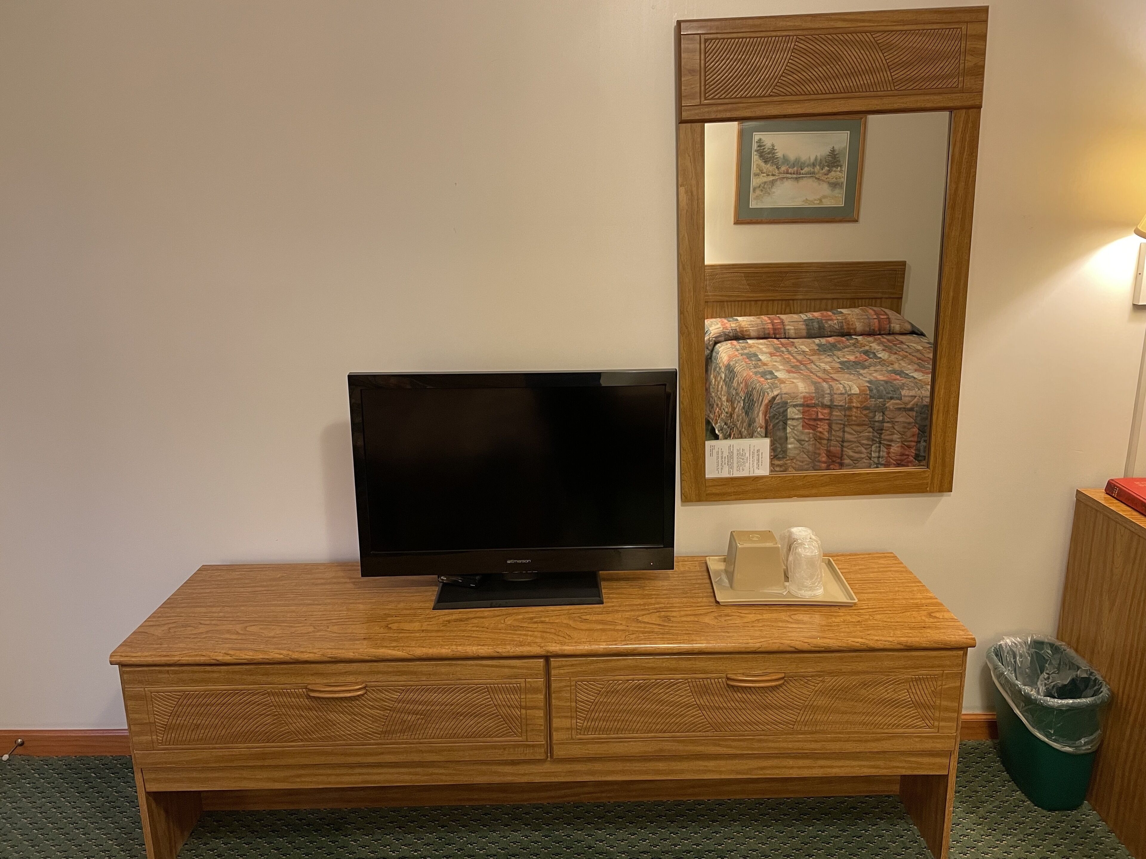 A wooden desk with a television, a framed picture above it, and a bed with a patterned bedspread in the reflection.