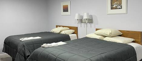 A hotel room with two beds, a wooden headboard, a nightstand, a wall-mounted lamp, and two framed pictures.