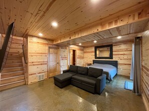 L’intérieur d’une cabane en rondins, avec un escalier en bois, un canapé d’angle, un lit et un grand miroir au mur.