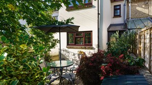 Une terrasse extérieure avec une table ronde et des chaises, entourée d’une végétation luxuriante et d’un petit jardin.