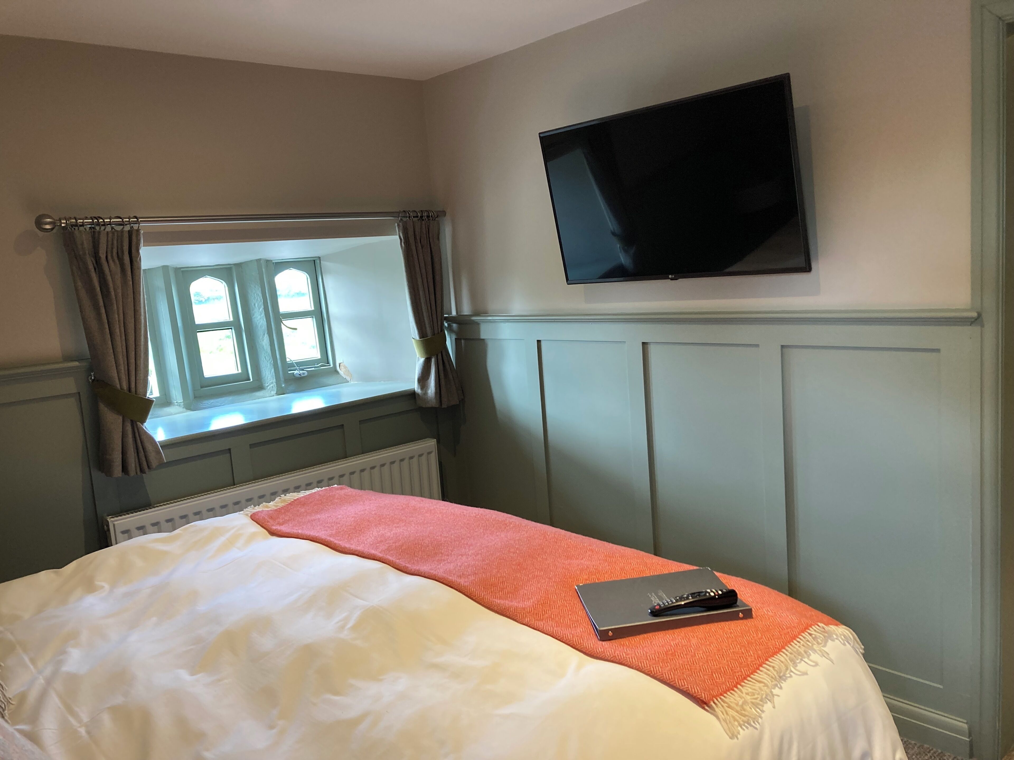 A bedroom with a bed, a window with curtains, a radiator, a television mounted on the wall, and a notebook on the bed.