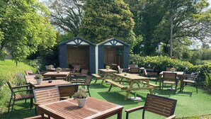 Un jardin avec des tables et des chaises en bois, deux petits abris et une variété de plantes.