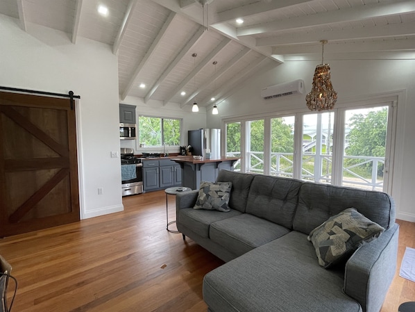 A spacious living area with a grey sofa, wooden flooring, and a kitchen in the background.