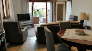 Een woonkamer met een televisie, bank, stoelen, een tafel met placemat, een lamp en een schuifpui naar een balkon.