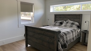 Une chambre à coucher avec un lit en bois, une fenêtre avec des stores, une table de chevet avec une lampe, et une vue sur les arbres à l’extérieur.