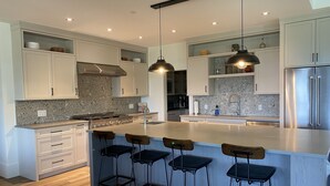 Une cuisine moderne avec des meubles blancs, un îlot central doté de tabourets de bar, des appareils en acier inoxydable et un éclairage suspendu.