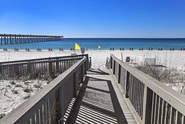 A beach with a wooden boardwalk leading to the water, a pier in the background, and beach chairs and umbrellas. - Newly remodeled and close to everything you need at Sea Glass Serenity. (Navarre)