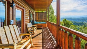 Una terraza de madera con asientos exteriores, vista a un paisaje boscoso y una cabaña de madera.